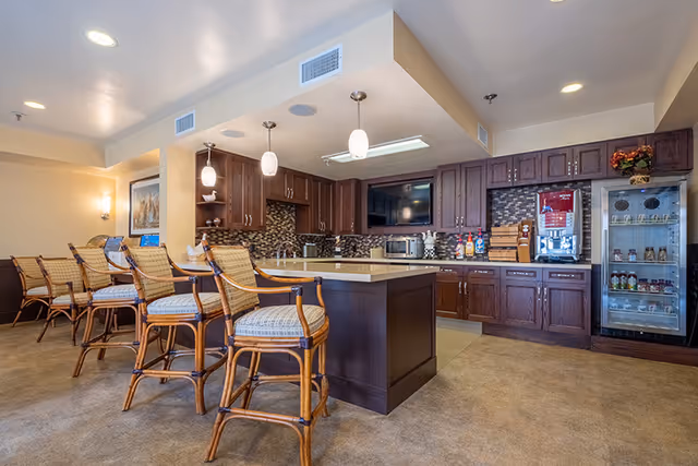 Interior view of a kitchen area in a senior living facility with a central island, four wooden bar stools with cushioned seats, dark wood cabinets, a tiled backsplash, a refrigerator with glass door stocked with beverages, and various kitchen appliances and condiments on the counters.