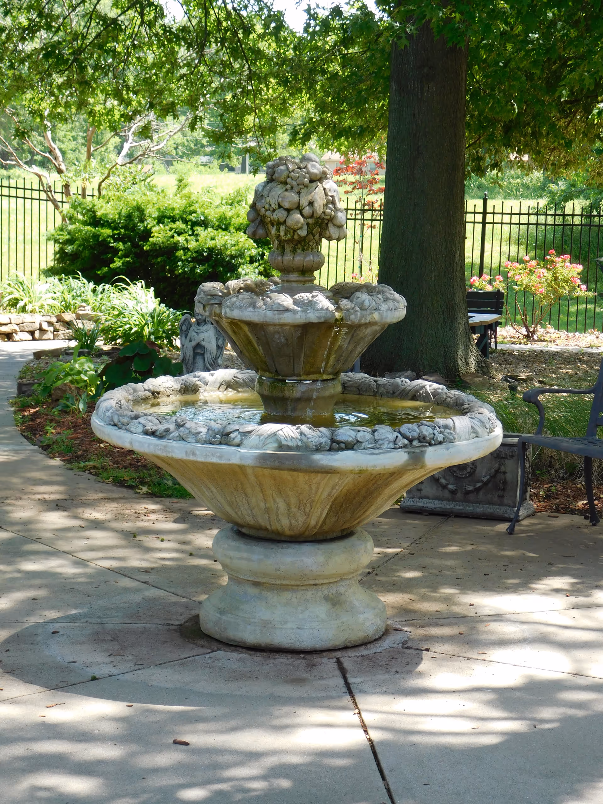 A decorative stone water fountain with a tiered design featuring carved fruit and floral motifs, situated on a paved area surrounded by greenery, trees, and garden benches in an outdoor garden setting.