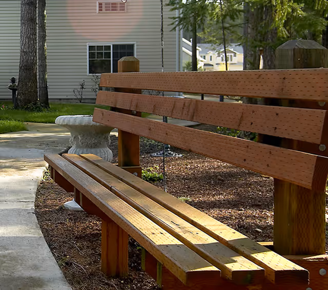 Wooden outdoor bench beside a concrete walkway in a landscaped courtyard with a building in the background.