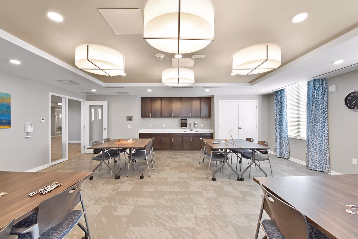 A bright, modern common room with several tables and chairs arranged for group activities. The room features large ceiling lights, a wall clock, blue patterned curtains on a window, and a cabinet with a countertop and sink along the back wall. The floor has a patterned tile design, and the walls are painted light gray.