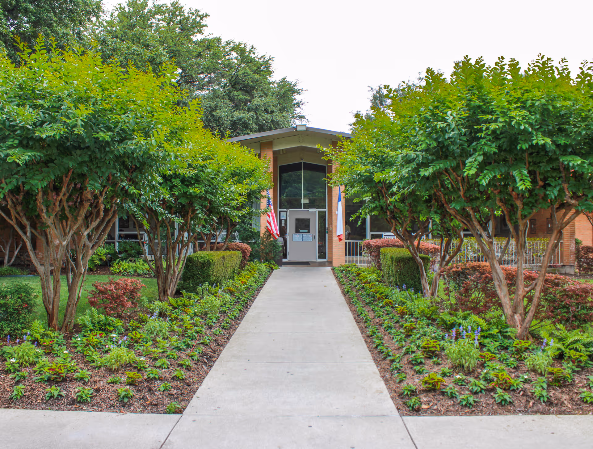Front entrance of a brick building with a concrete walkway leading up to the door, flanked by neatly trimmed bushes and small trees on both sides. Two flags are displayed near the entrance, and the area is surrounded by greenery and landscaping.