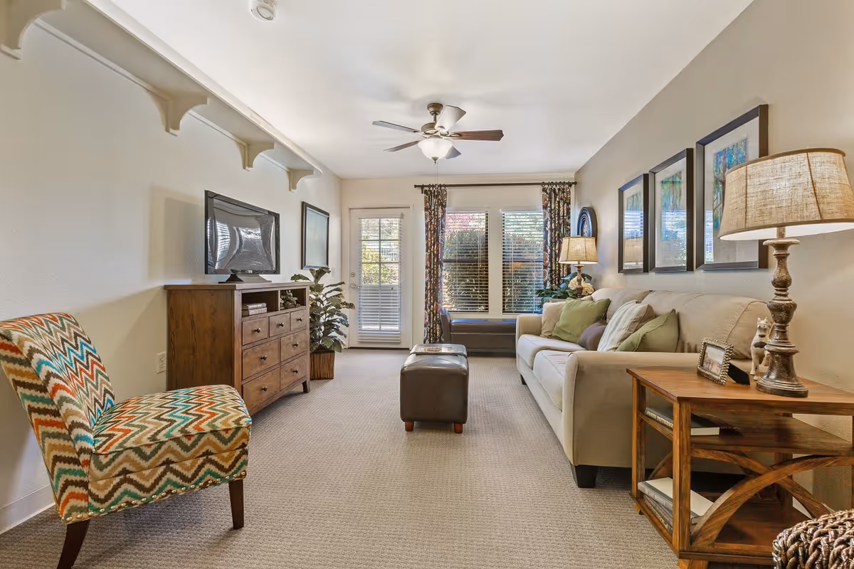 A cozy living room with a beige sofa adorned with green and brown pillows, a wooden side table with a lamp and picture frame, a patterned accent chair, a wooden dresser with a flat-screen TV on top, and a ceiling fan. The room has large windows with floral curtains and a glass door leading outside.