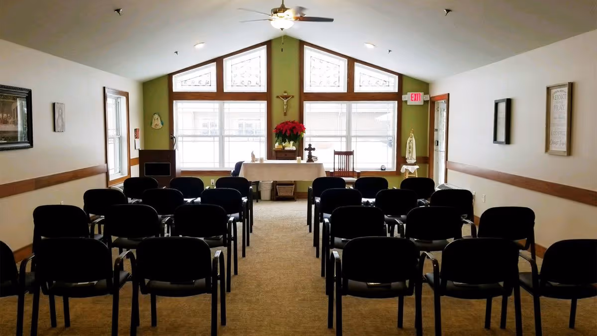 Small chapel-like meeting room with rows of chairs facing a table altar, lectern, and large windows with triangular transom panes.