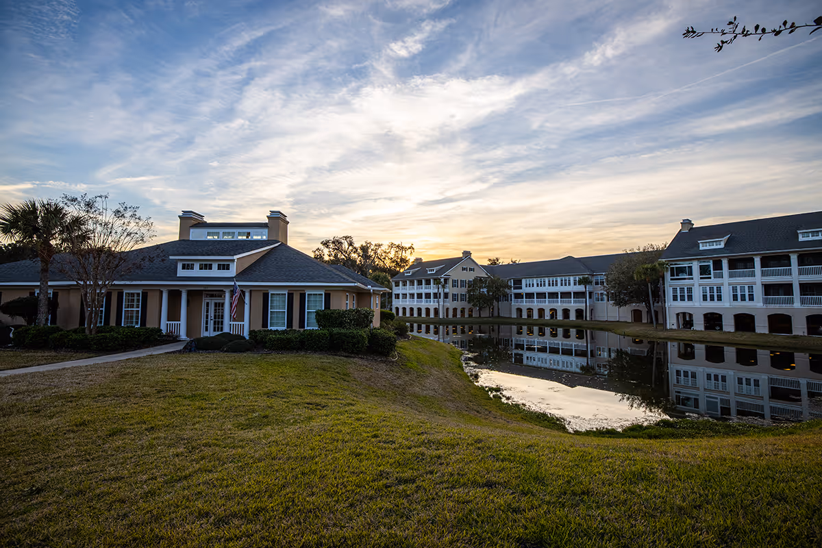 Exterior view of Marsh's Edge senior living facility at sunset, showing multiple buildings with white and beige walls, large windows, and a small pond reflecting the buildings and sky. The foreground features a grassy area and a pathway leading to one of the buildings.