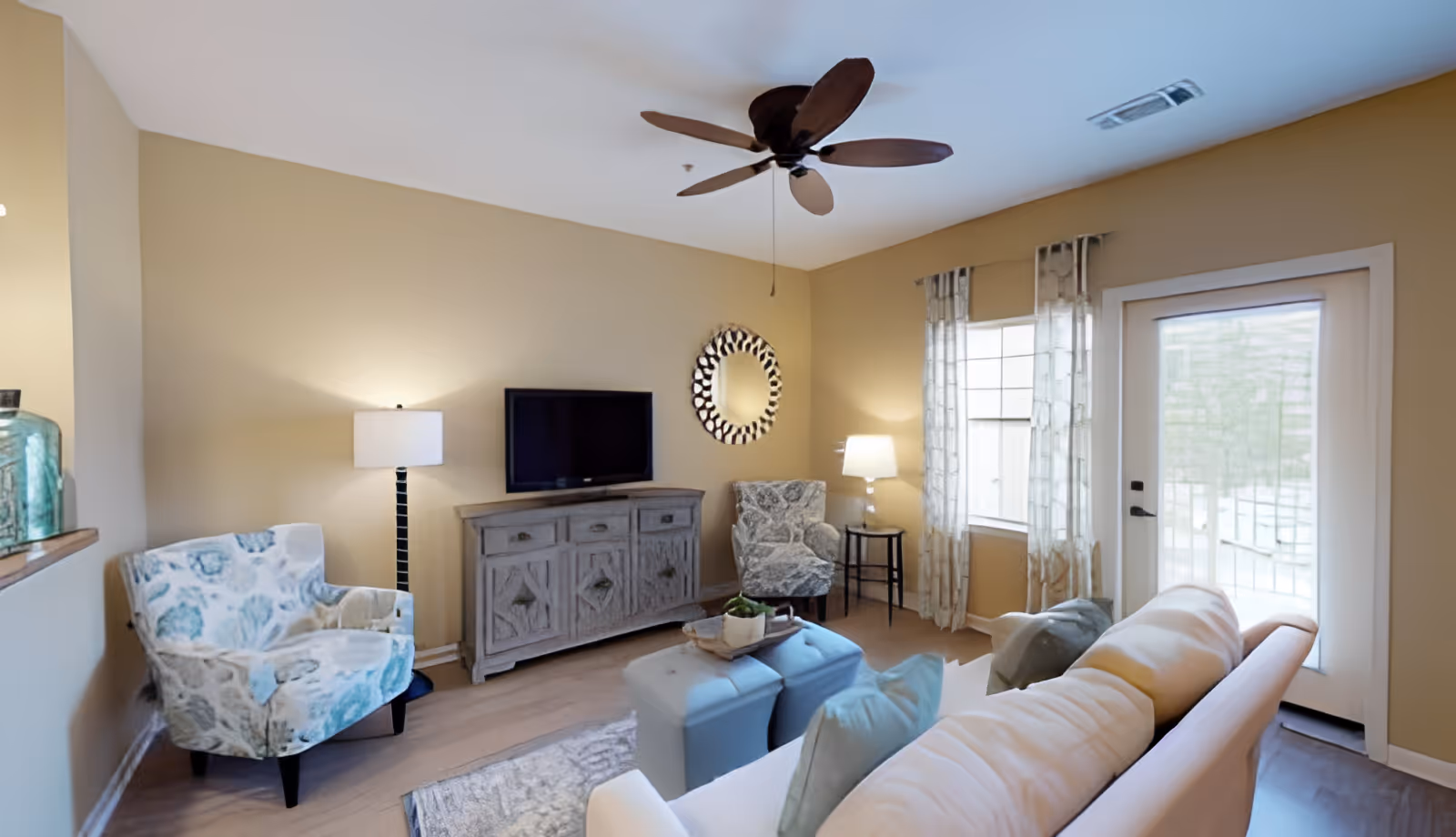 A cozy living room with beige walls, a ceiling fan, a flat-screen TV on a rustic wooden console, two patterned armchairs, a white sofa with cushions, two blue ottomans, a small side table with a lamp, and a glass door with curtains letting in natural light.