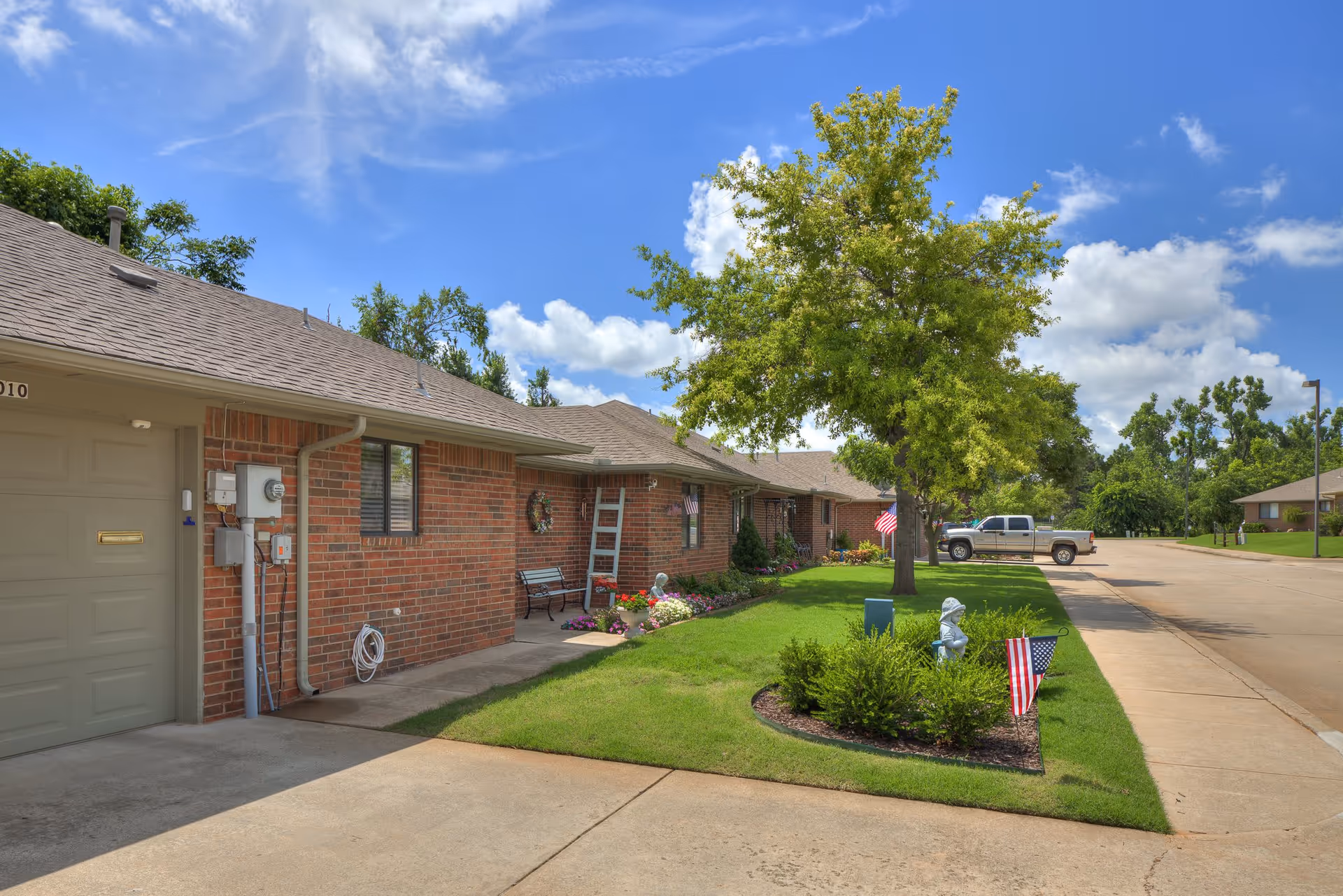 Exterior view of a single-story brick residential building with a well-maintained lawn, a tree, flower beds, and American flags. A pickup truck is parked on the street beside the building under a partly cloudy blue sky.
