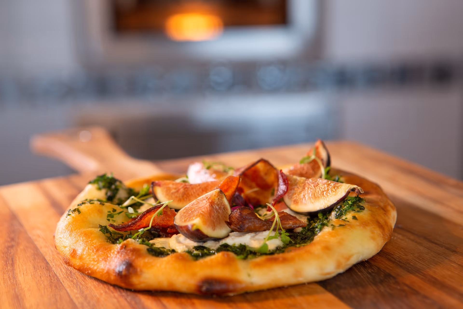 Close-up of a freshly baked flatbread pizza topped with figs, greens, and crispy bacon on a wooden cutting board in a kitchen setting.