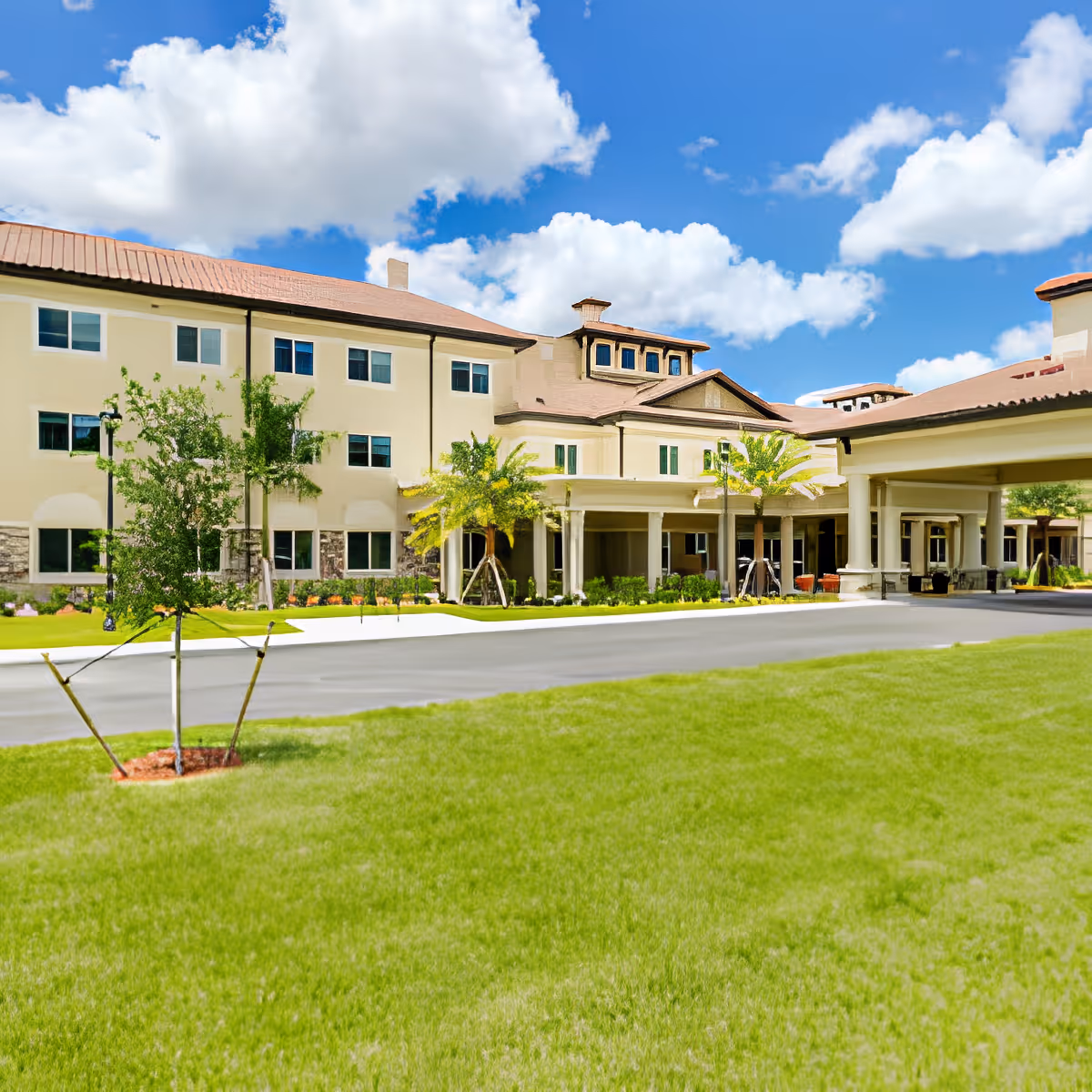 Exterior view of a large senior living facility building with beige walls and a red-tiled roof under a blue sky with scattered clouds. The building is surrounded by green grass, small trees, and a paved driveway leading to a covered entrance.