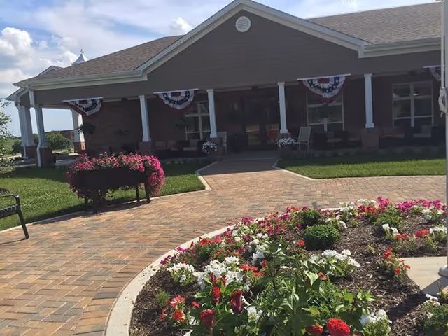 Front entrance of a senior living building with a covered porch, brick walkway, and flower beds in the foreground.