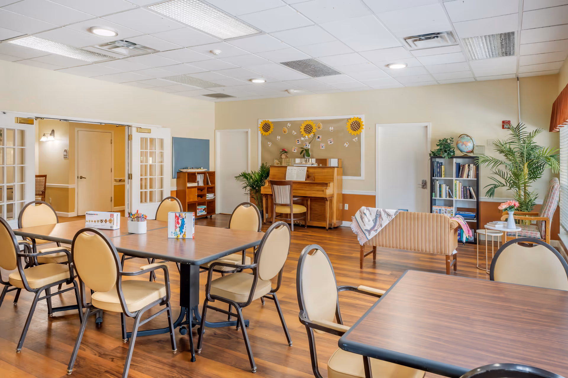 A bright and spacious common area with multiple tables and chairs arranged for group activities or dining. The room features a piano against the far wall, a bookshelf filled with books, a small couch with a blanket draped over it, and several plants adding greenery. The walls are light-colored, and the floor is wooden. There are double glass doors on the left side and a large window with curtains on the right.