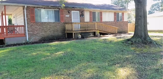 Single-story brick building with a wooden ramp and steps leading to the front door, surrounded by a grassy yard and a large tree on the right side.