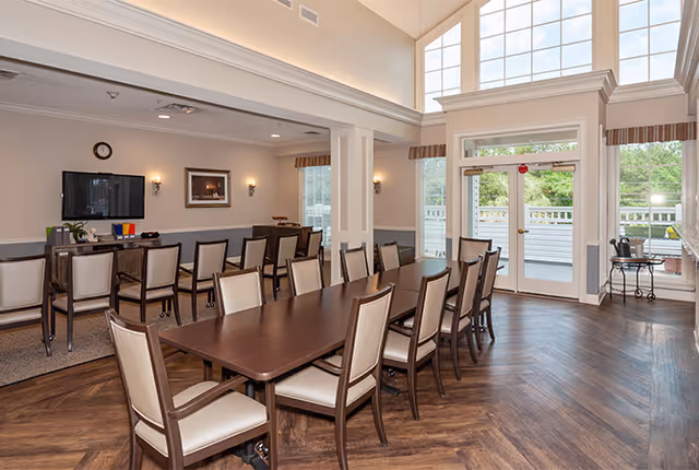 Bright communal dining room with long wooden tables, upholstered chairs, a wall-mounted TV, and large windows with double doors to an outdoor deck.