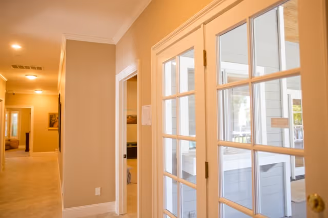 Interior view of a hallway in a senior living facility with beige walls and carpeted floor. On the right side, there are white-framed glass double doors leading to another room with large windows. The hallway extends to the left with doorways and framed pictures on the walls.
