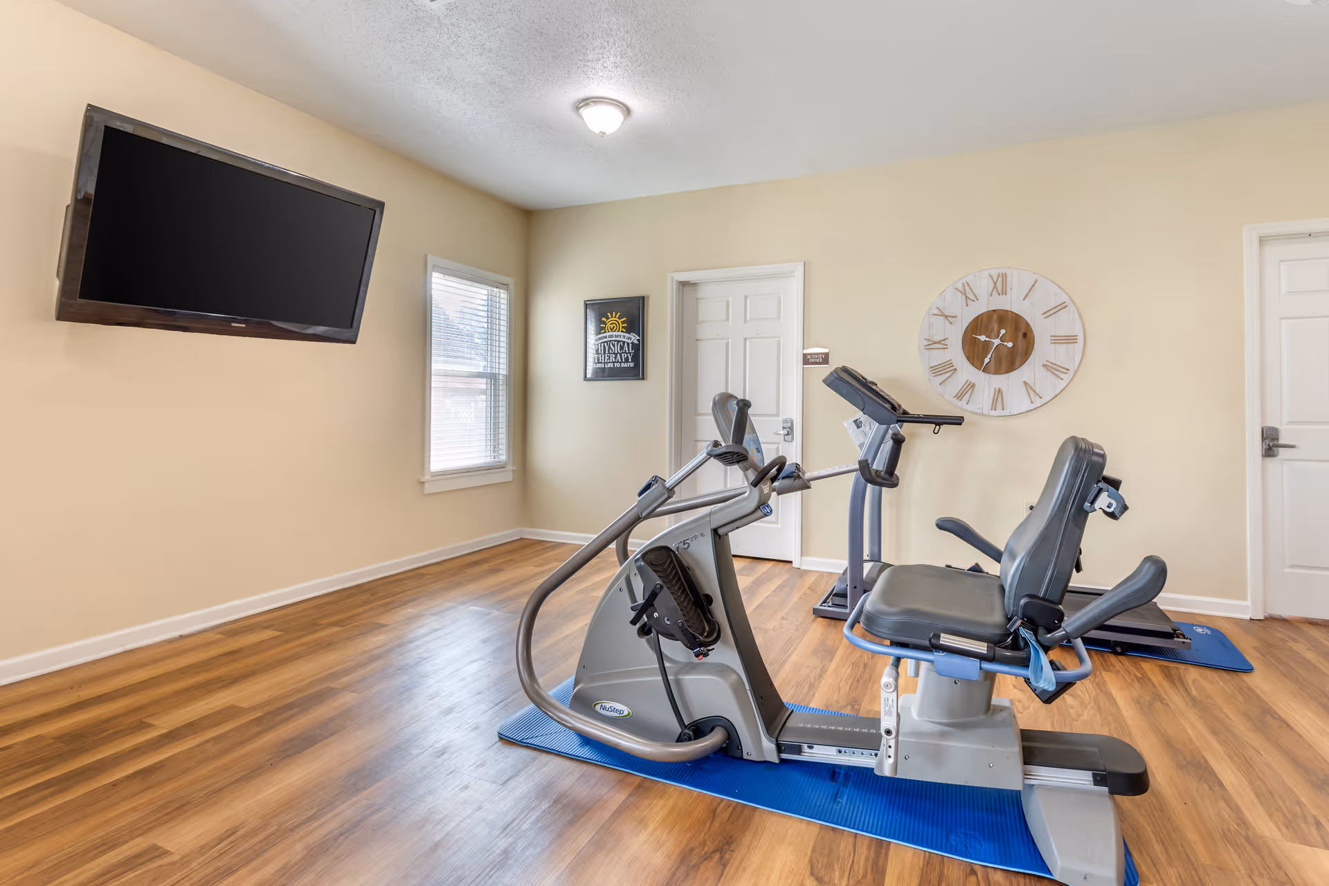 A small exercise room with wooden flooring featuring a recumbent exercise bike and a treadmill on blue mats. The room has beige walls, a large wall clock with Roman numerals, a window with blinds, a wall-mounted flat-screen TV, and two white doors, one labeled 'Activity Office'.