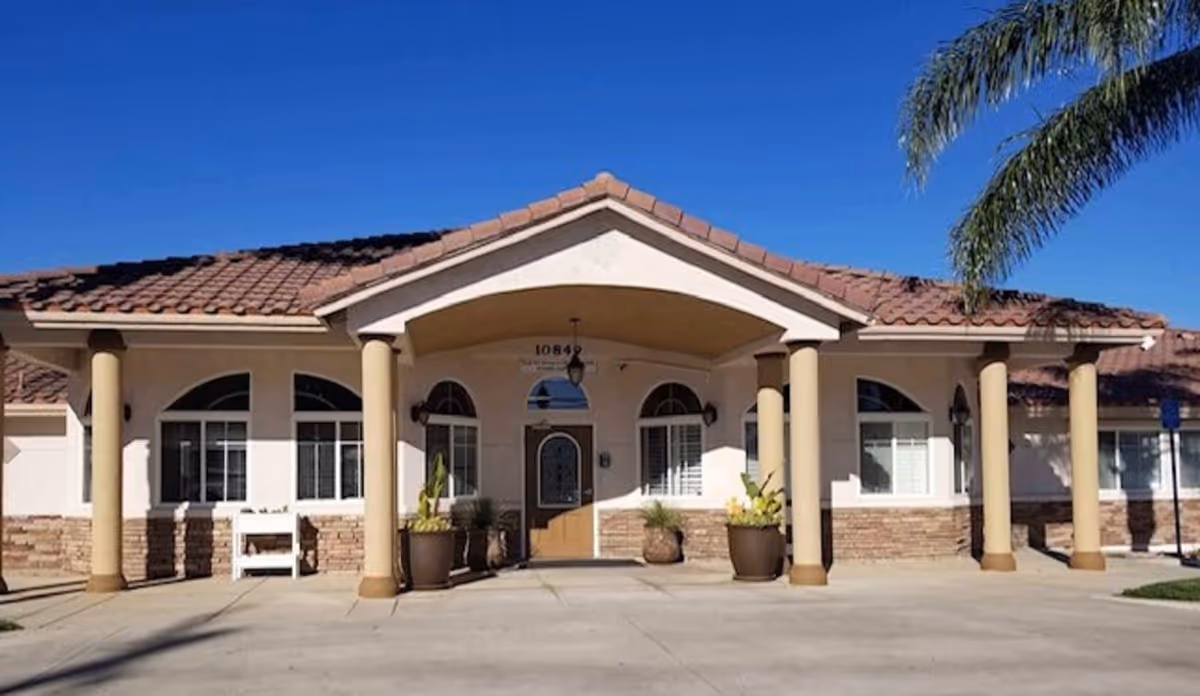 Front exterior view of a single-story building with a tiled roof and beige walls, featuring a covered entrance supported by columns, potted plants near the doorway, and several windows with white shutters under a clear blue sky.