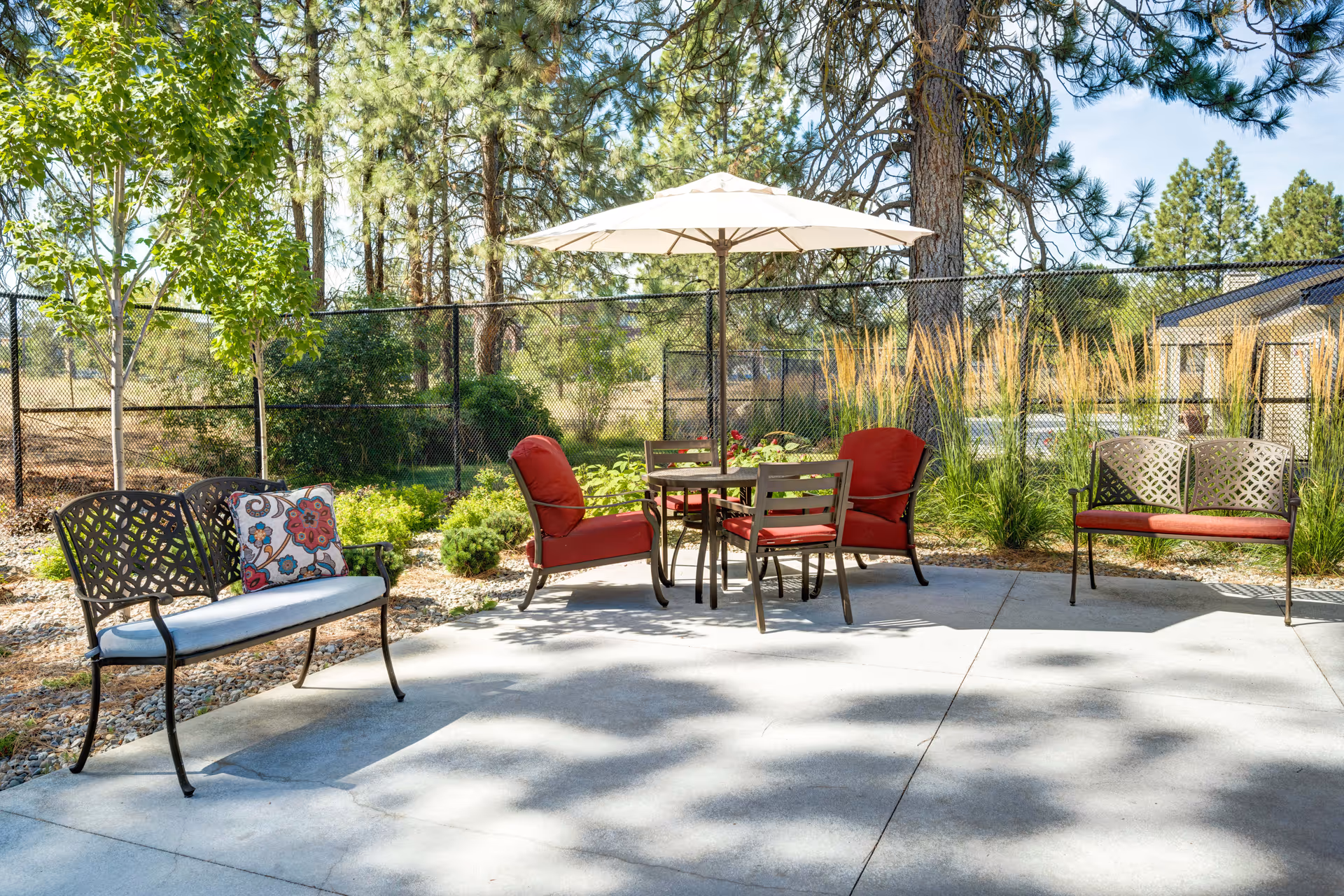 Outdoor patio area with metal benches and chairs with red cushions around a table with a white umbrella. The patio is surrounded by trees, shrubs, and a black chain-link fence under a clear blue sky.