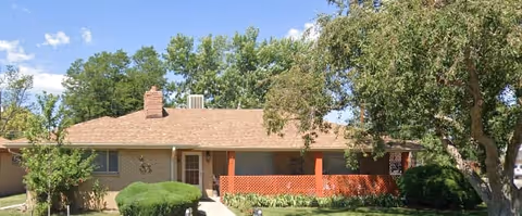 Single-story brick residential building with a brown shingled roof, a covered porch with red lattice railing, surrounded by green bushes and trees under a blue sky with some clouds.