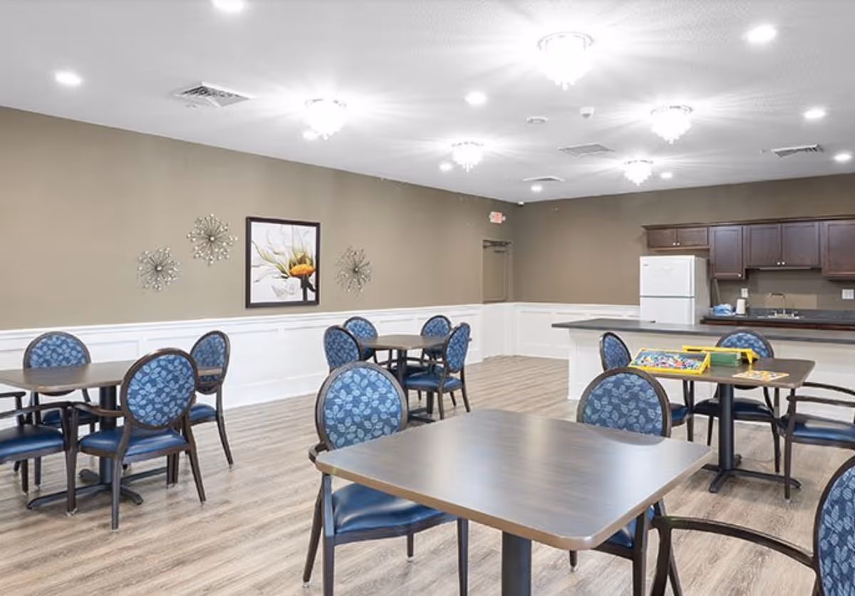 A bright and spacious dining area with several tables and blue cushioned chairs. The room features light wood flooring, beige walls with white wainscoting, and decorative wall art. In the background, there is a kitchenette with dark wood cabinets, a white refrigerator, and a countertop. The ceiling has multiple light fixtures providing ample lighting.