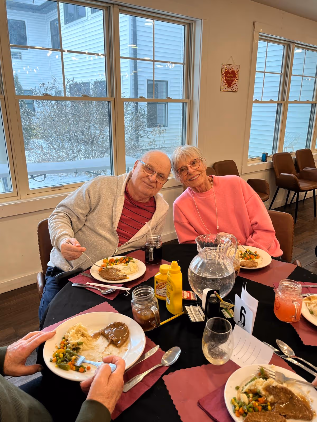 Two elderly people sitting at a round dining table with plates of food including mashed potatoes, vegetables, and meat with gravy. The table has condiments, a pitcher of water, glasses, and utensils. Large windows show an outside view of a building and some bushes.