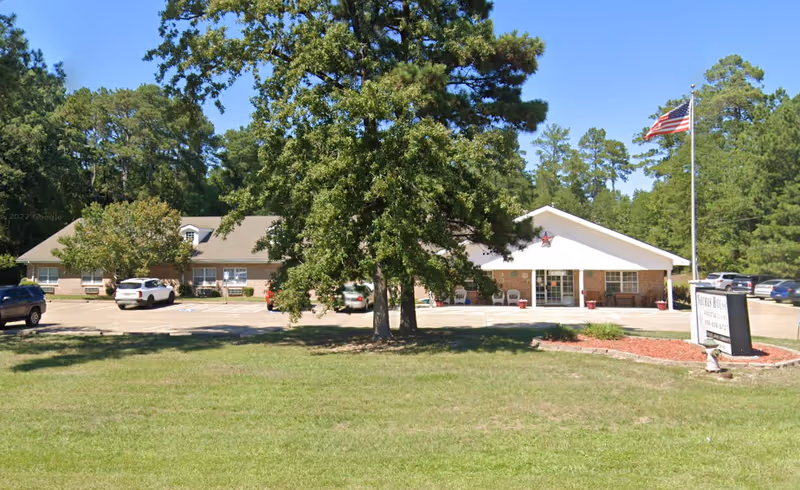 Exterior view of Countryside Senior Living of Lufkin, a single-story brick building with a white entrance canopy, surrounded by trees and a grassy lawn. Several cars are parked in the parking lot in front of the building, and an American flag is flying on a flagpole near the entrance.