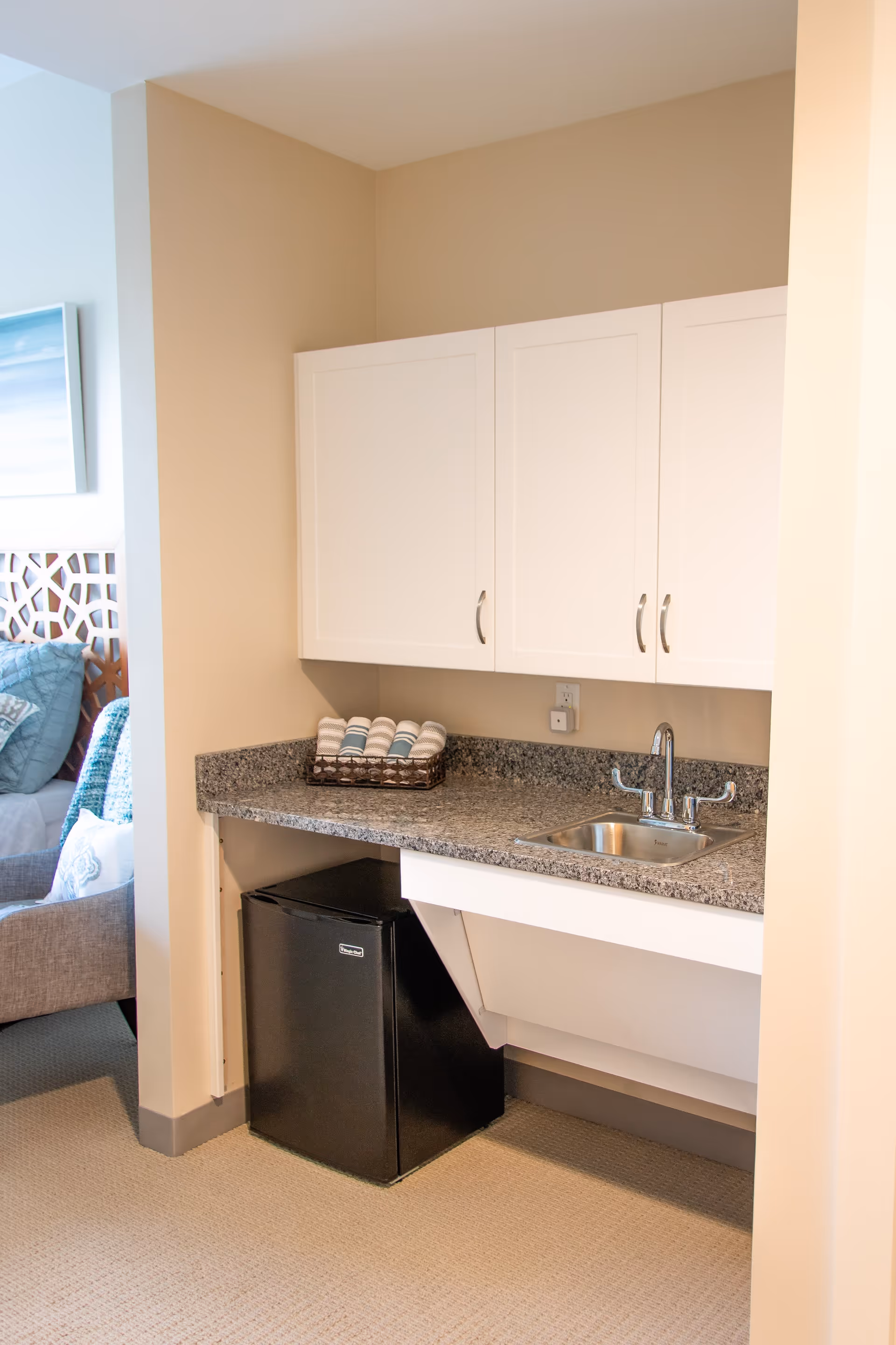 A small kitchenette area with a granite countertop, a stainless steel sink with a faucet, white cabinets above, a black mini refrigerator underneath, and a basket with rolled towels on the counter. Part of a living room with a gray armchair and a bed with blue pillows is visible to the left.