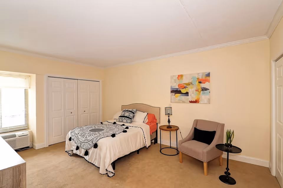 A neatly arranged bedroom with a single bed covered in white and black patterned bedding, a beige upholstered chair with a black cushion, two small round side tables, one with a lamp and the other with a small plant, a colorful abstract painting on the wall, a window with blinds and an air conditioning unit below it, and a double-door closet.