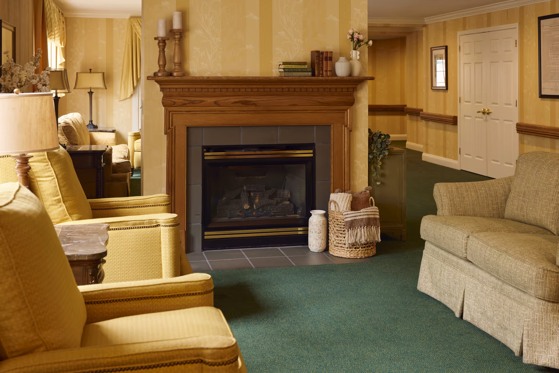 A cozy living room area with a wooden fireplace in the center, flanked by yellow upholstered armchairs on the left and a green upholstered sofa on the right. The room features green carpet, yellow patterned wallpaper, decorative candles, books, and vases on the fireplace mantel, and a basket with blankets on the floor next to the fireplace. A hallway with framed pictures and double doors is visible in the background.