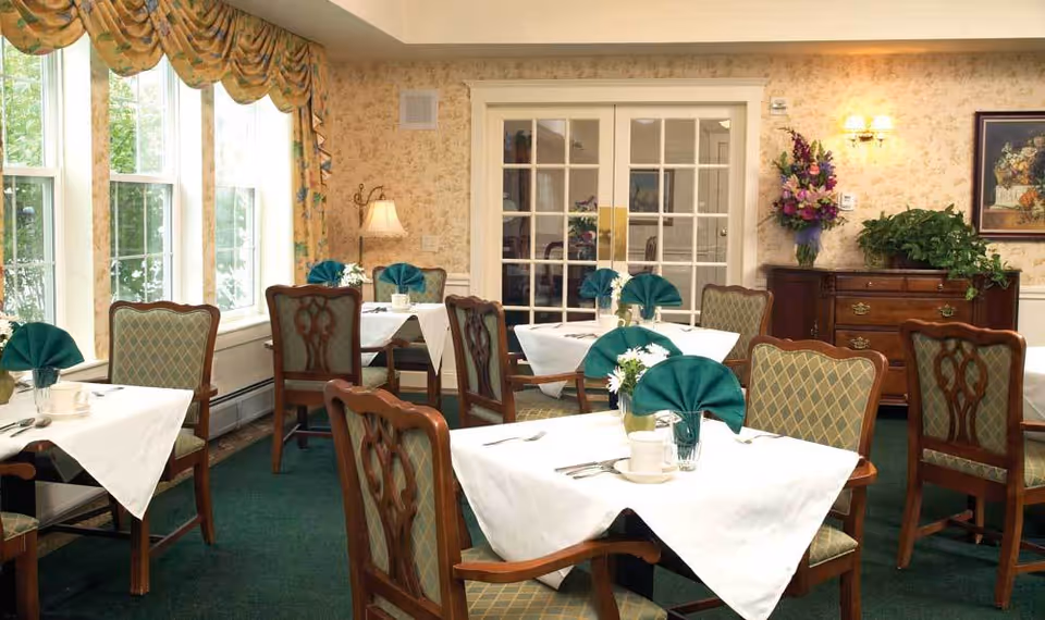 A dining room with several tables covered in white tablecloths, each set with green folded napkins, cups, and silverware. The room has large windows with patterned curtains, green carpet, and wooden chairs with upholstered seats and backs. A wooden sideboard with plants and a floral arrangement is against the wall, which is decorated with wallpaper and a framed painting.