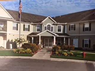 Two-story beige senior living facility entrance with columns, landscaped flower beds, an American flag, and a walkway leading to the front doors.