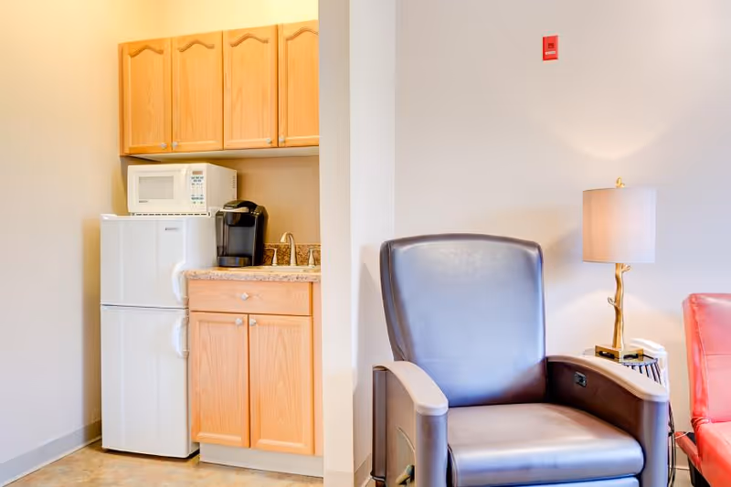A small kitchenette area with wooden cabinets, a white microwave on top of a white mini refrigerator, and a black coffee maker on the countertop. Next to the kitchenette is a dark brown leather armchair and a side table with a lamp and a telephone.