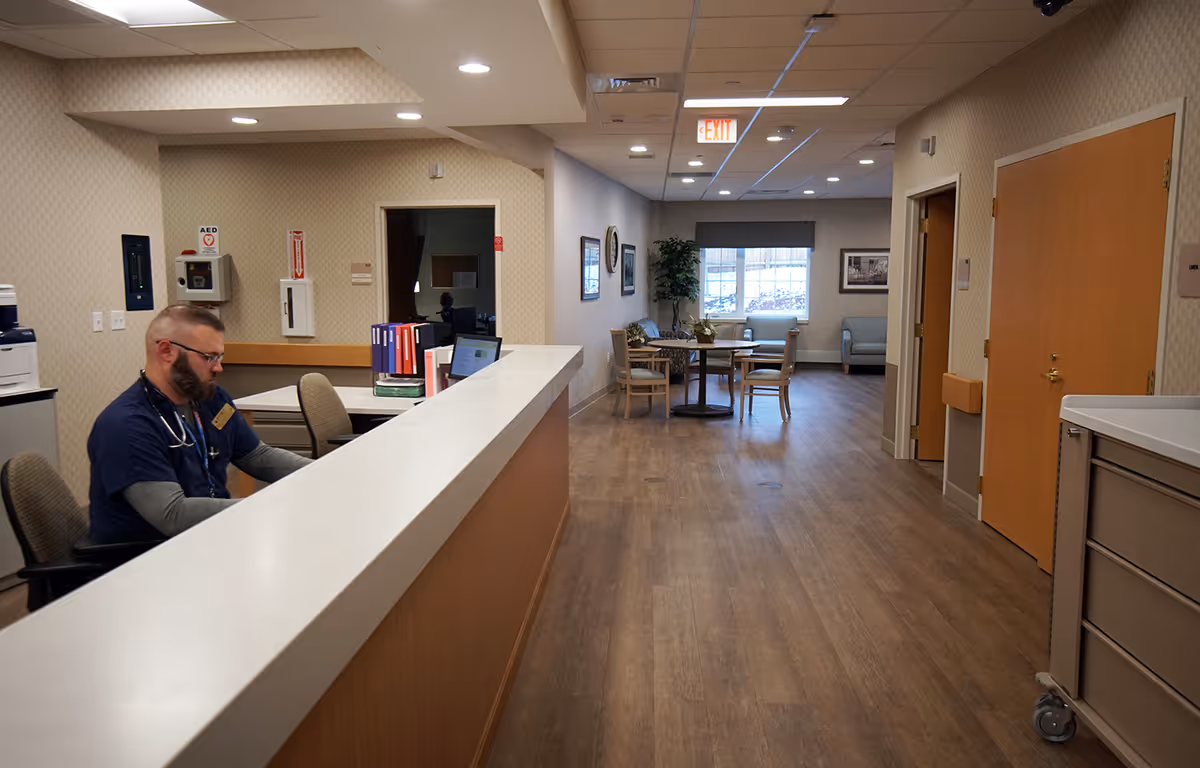 Reception desk with a seated staff member on the left and a long hallway leading to a seating area with tables and chairs.