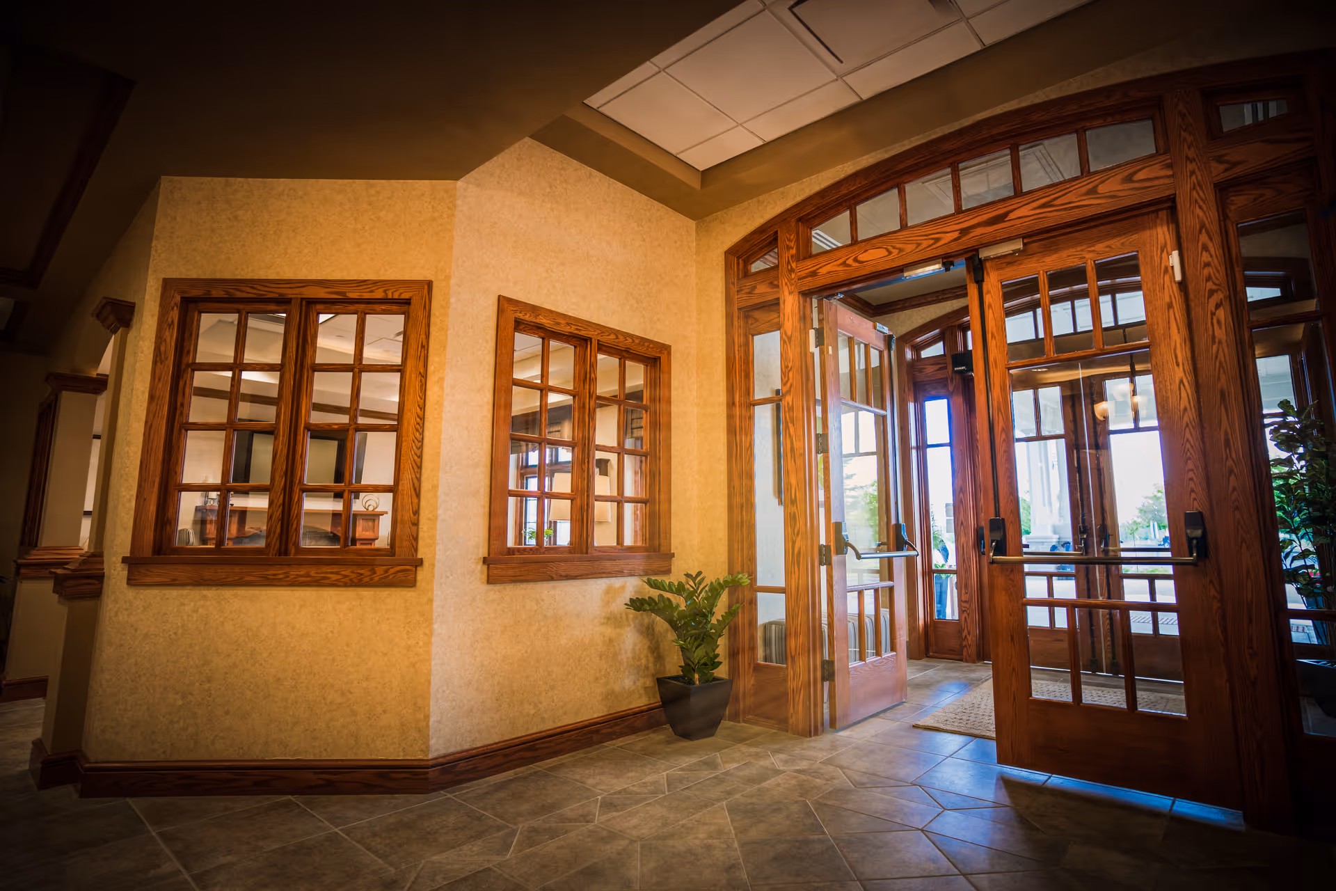 Interior view of an entrance area with large wooden-framed glass doors and windows. The floor is tiled, and there is a potted plant near the corner wall. Natural light is coming through the glass doors, illuminating the space.