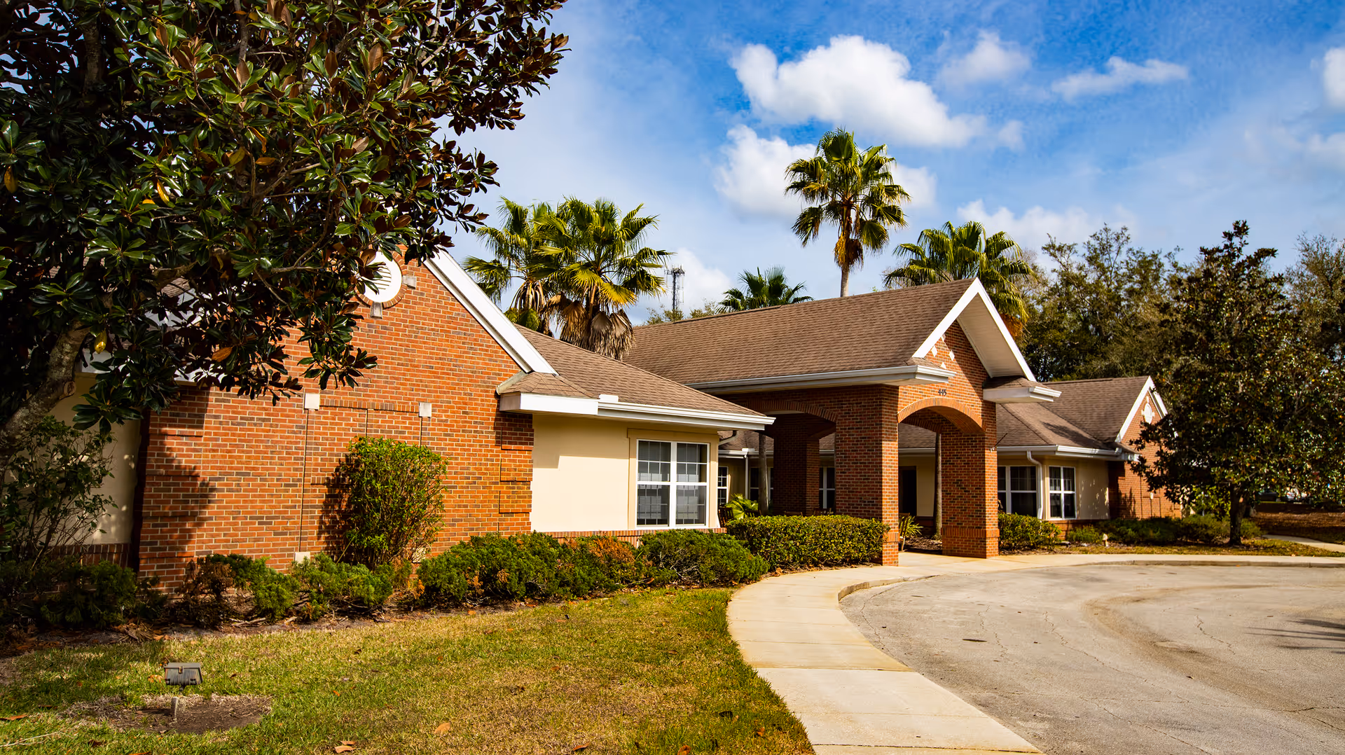 Exterior view of a single-story brick building with a covered entrance, surrounded by palm trees and other greenery under a partly cloudy sky.
