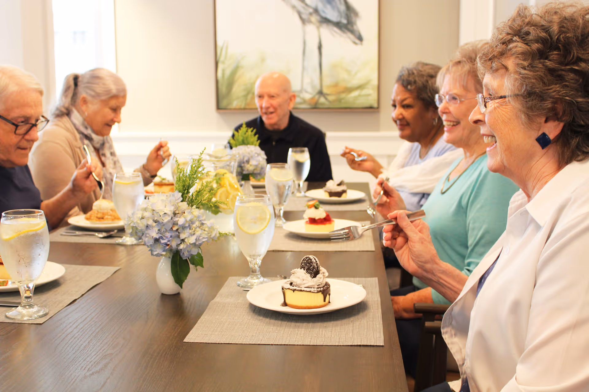 A group of six elderly people sitting around a dining table enjoying dessert and water with lemon. The table is decorated with a vase of blue hydrangeas and a pitcher of lemon water. The room has light-colored walls and a framed painting of a bird in the background.