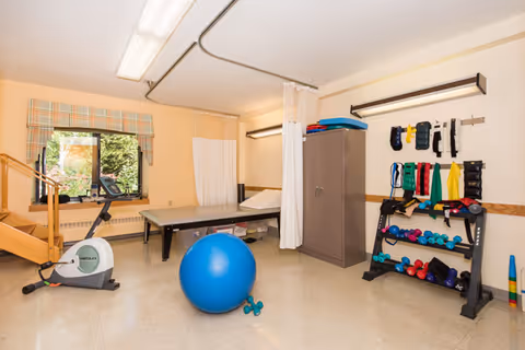 A rehabilitation room with exercise equipment including a stationary bike, a large blue exercise ball, dumbbells, resistance bands, and a padded therapy table. The room has a window with a plaid valance, beige walls, and a storage cabinet. There are ceiling-mounted curtains for privacy.