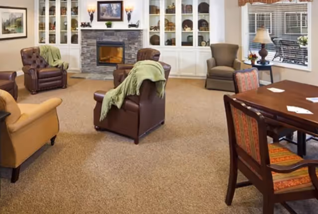 A cozy living room area in a memory care community featuring several armchairs with green throws, a stone fireplace with a framed picture above it, built-in white shelves with decorative items, a wooden table with chairs, a table lamp on a side table, and a window with blinds.