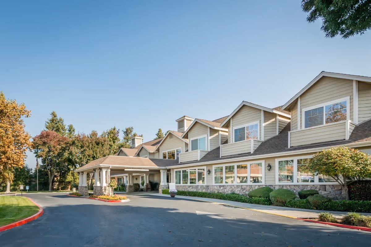 Front exterior of a two-story senior living facility with a covered entrance, landscaped shrubs, and a paved driveway.