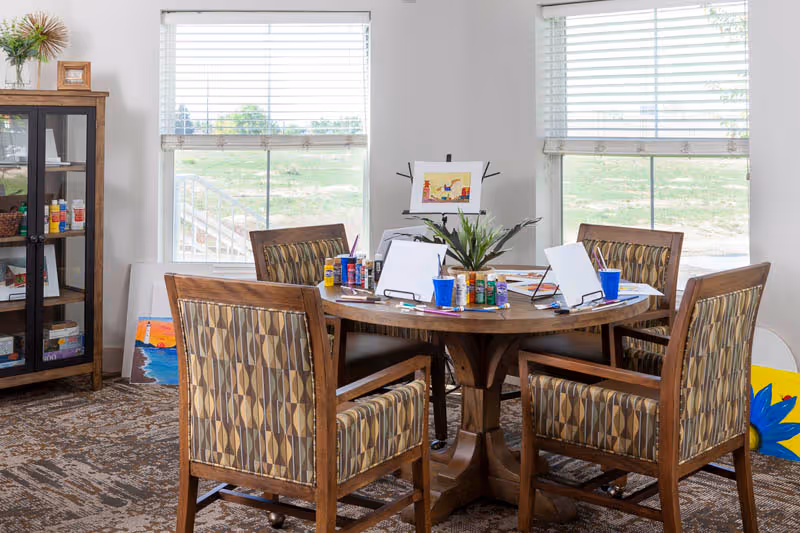 A round wooden table with four patterned cushioned chairs around it in a bright room with two large windows. The table is set up for painting with canvases on stands, paint bottles, brushes, and cups. A small plant is in the center of the table. To the left, there is a glass-front cabinet with art supplies and a framed picture on top. The floor is carpeted with a patterned rug.