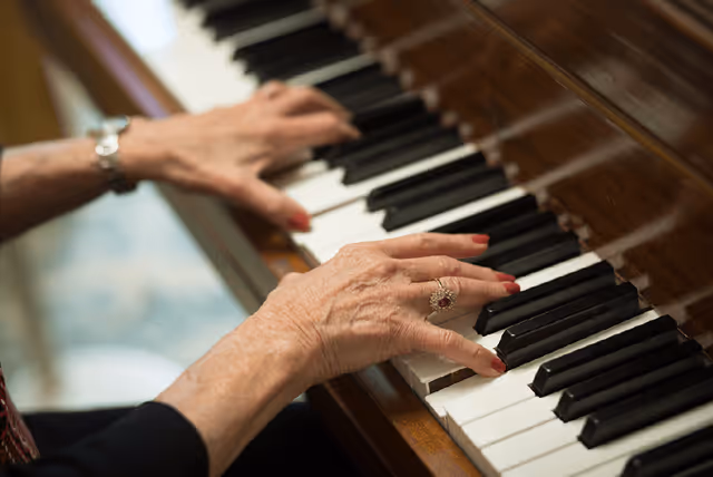 Close-up of an elderly person's hands playing a wooden piano keyboard, showing detailed wrinkles and a ring on one finger.