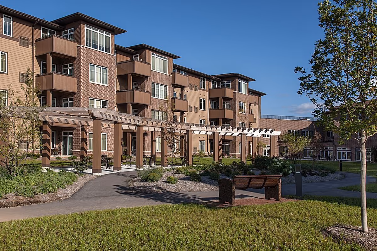 Outdoor view of a senior living facility named Prairie Gate featuring a multi-story building with balconies, a pergola walkway, landscaped garden areas, trees, and a bench under a clear blue sky.