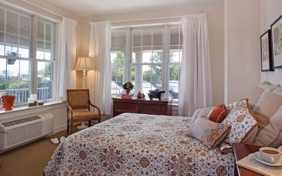 A cozy bedroom with a large bed covered in a patterned quilt and multiple pillows. There is a wooden chair with a patterned cushion next to a floor lamp by the window. The room has large windows with white curtains letting in natural light. A wooden dresser with decorative items and books is placed under the windows. A cup of coffee and a book are on a bedside table.