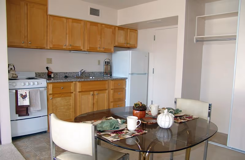 A small kitchen area with light wood cabinets, a white refrigerator, and a white stove with a rooster-themed towel hanging on the handle. In front of the kitchen is a glass-top dining table set with placemats, cups, and decorative pumpkins. Two cream-colored chairs are positioned at the table. To the right, there is an open closet space with a hanging rod and shelf.