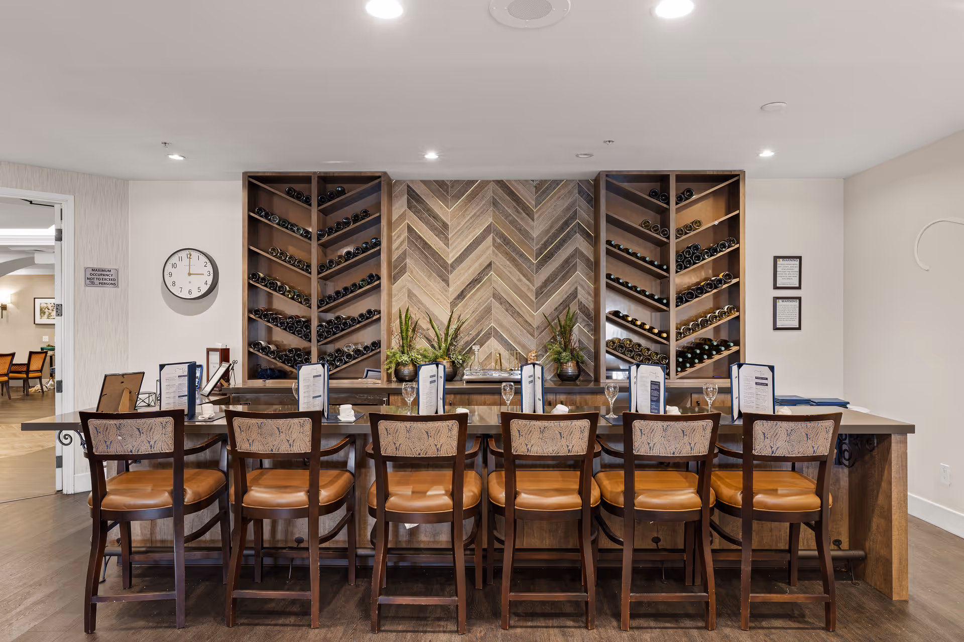 Bar-style dining counter with eight wooden chairs in front of built-in wine racks and a chevron wood accent wall.