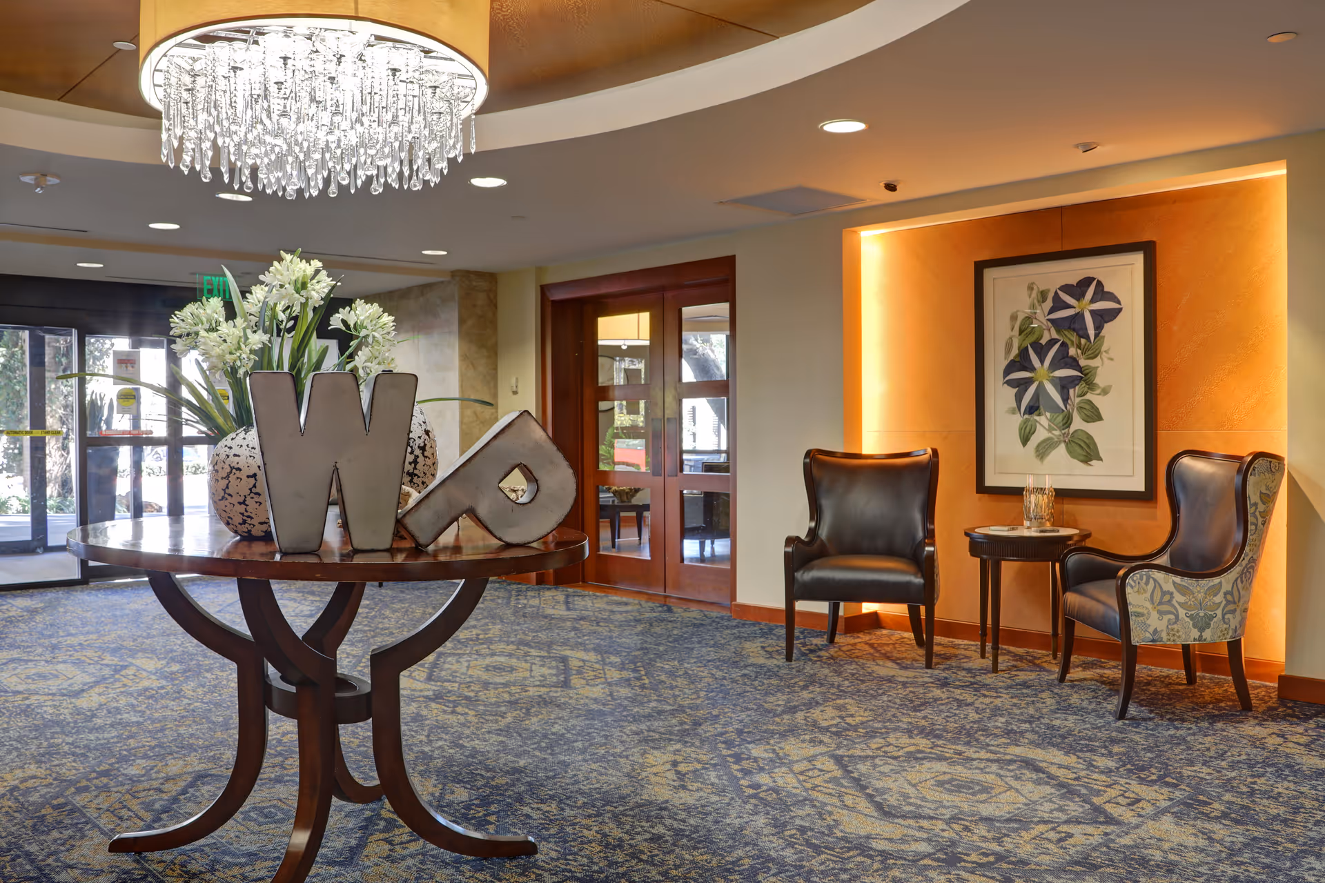 Welcoming lobby with a round table displaying decorative letters and flowers, a crystal chandelier overhead, and seating against an orange accent wall.