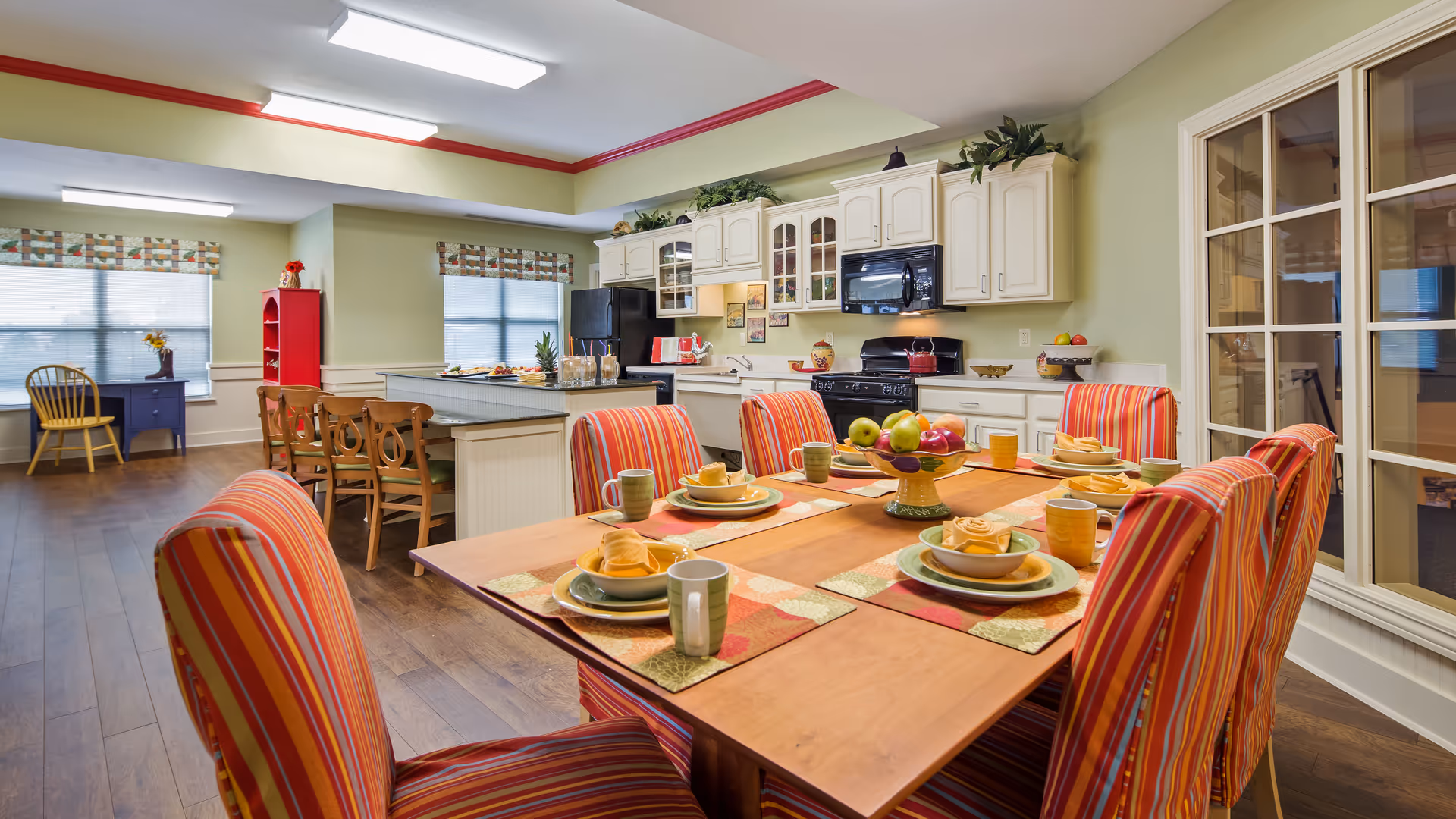 A bright and spacious kitchen and dining area in a senior living facility. The dining table is set with six place settings, each with plates, bowls, cups, and yellow napkins. The chairs around the table have red and orange striped covers. The kitchen features white cabinets, a black refrigerator, stove, and microwave. There is a kitchen island with wooden chairs and a fruit centerpiece on the dining table. The room has large windows with patterned valances and wooden flooring.