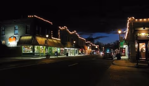 A nighttime street scene showing a row of buildings decorated with string lights along the rooflines. The street is dimly lit with a few street lamps, and some storefronts are visible with illuminated windows. The sky is dark with a hint of blue near the horizon.