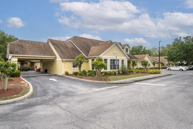 Exterior view of a single-story yellow building with a brown shingled roof, surrounded by small trees and landscaped bushes. There is a driveway and parking area with several cars parked, under a partly cloudy sky.