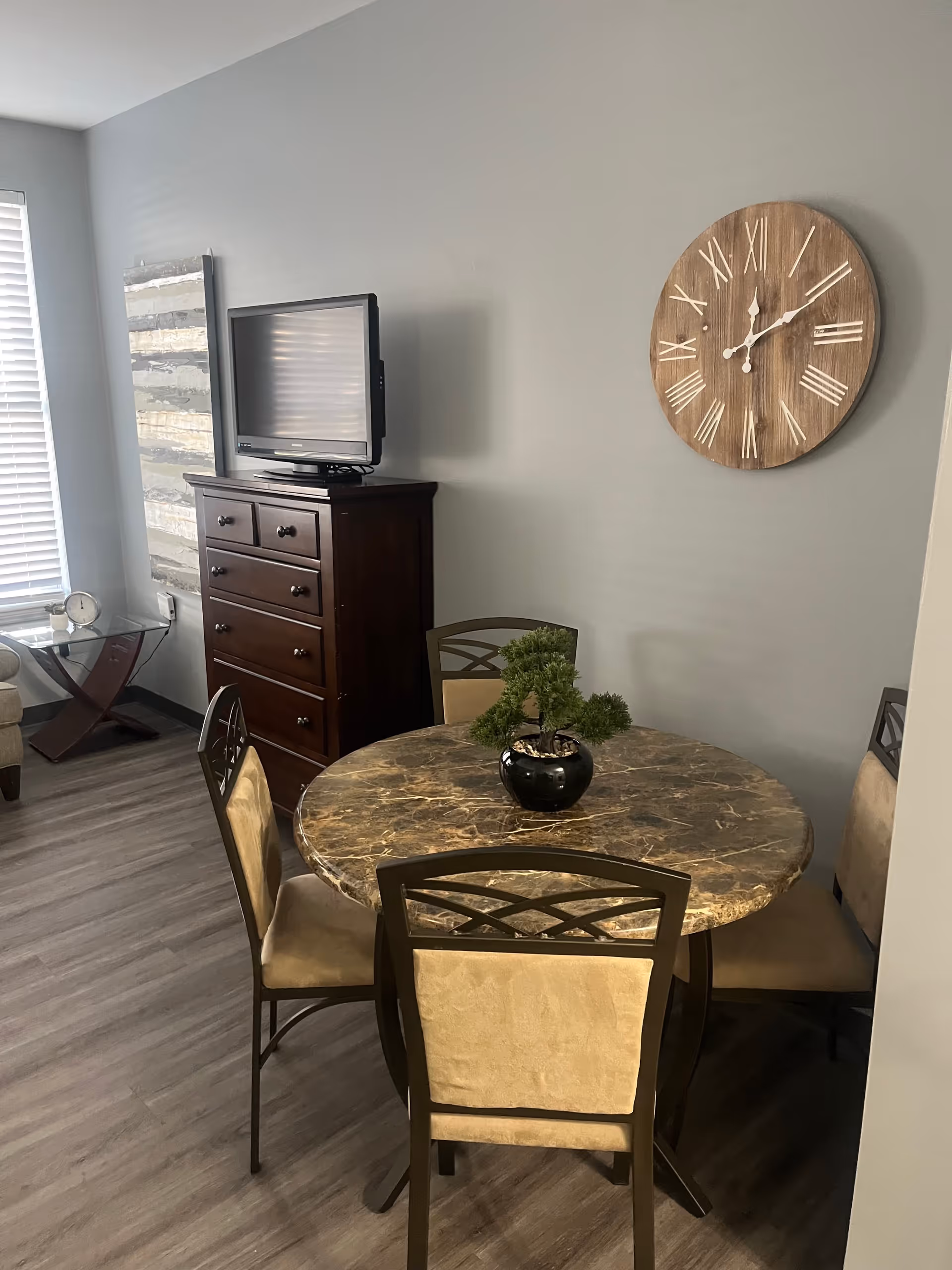 A small dining area with a round marble table surrounded by four cushioned chairs. A small potted plant is placed in the center of the table. On the wall above the table is a large wooden clock with Roman numerals. To the left, there is a dark wooden dresser with a flat-screen TV on top. A window with blinds and a small side table with a clock are visible in the background.
