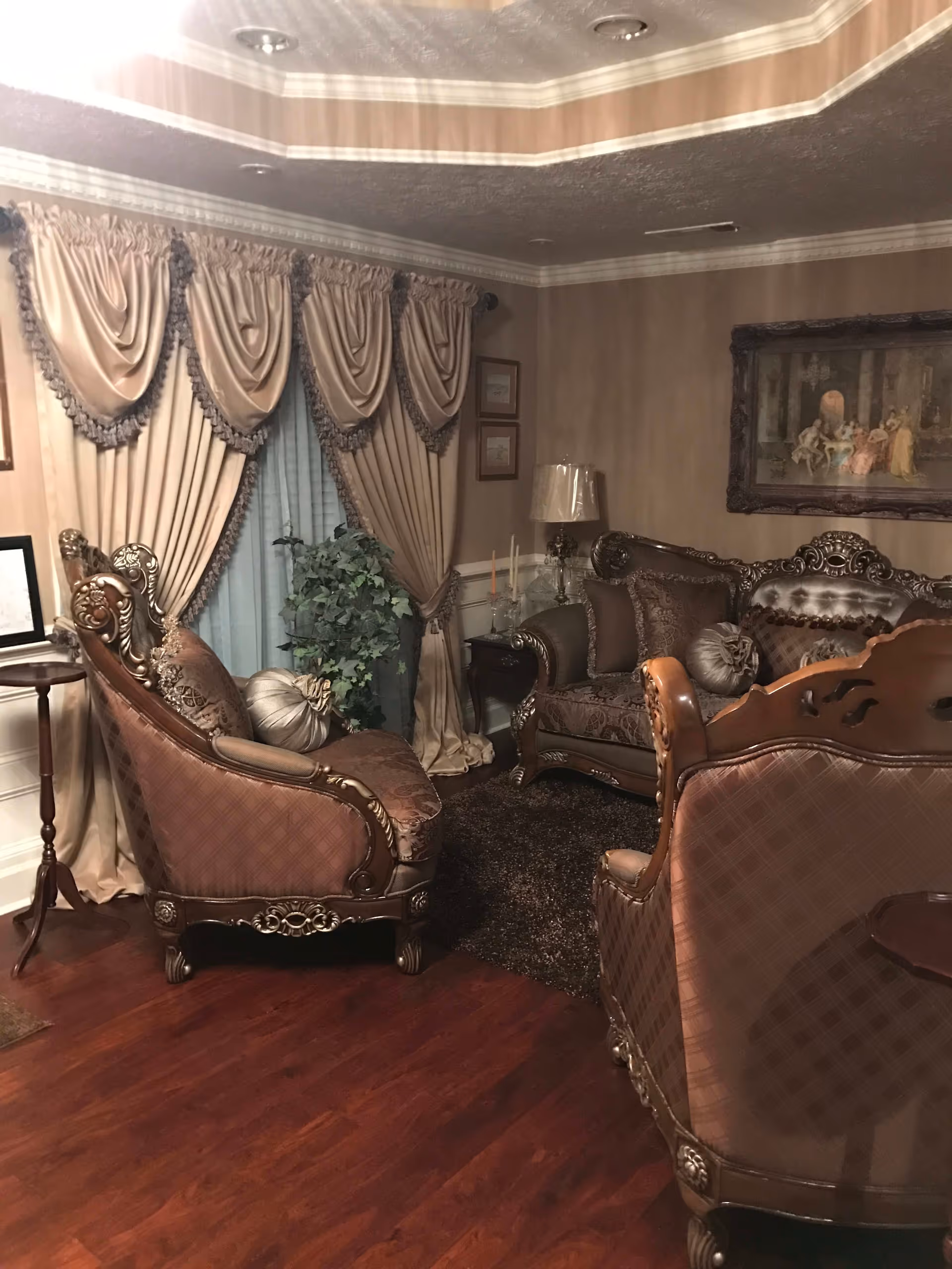 A cozy living room with ornate, vintage-style upholstered furniture including a sofa and armchairs. The room features heavy draped curtains with tassels, a small side table with a lamp, framed artwork on the walls, a potted plant near the window, and a dark area rug on polished wooden floors.