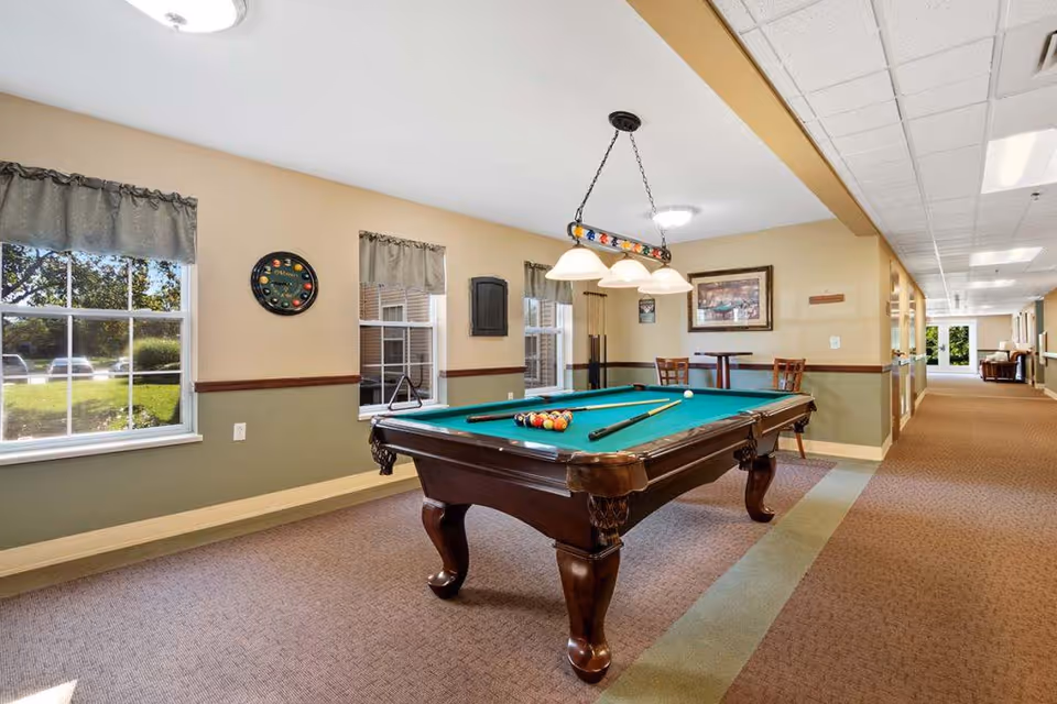 A well-lit indoor recreational room featuring a classic wooden pool table with green felt, pool cues, and billiard balls arranged for a game. The room has beige and green walls with two windows showing a green outdoor area. There is a hanging light fixture above the pool table, a small table with two chairs in the background, and a long carpeted hallway extending to the right.